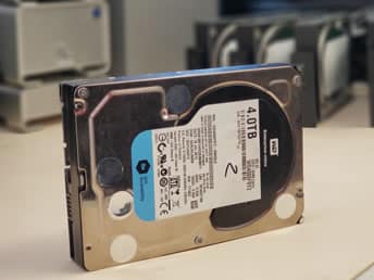 Hard Drive Data Recovery A 4TB internal hard drive stands upright on a desk, with blurred data recovery equipment in the background. The setting suggests a data recovery lab environment.