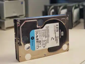 Hard Drive Data Recovery A 4TB internal hard drive stands upright on a desk, with blurred data recovery equipment in the background. The setting suggests a data recovery lab environment.