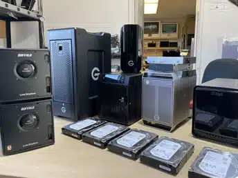 RAID Data Recovery Service A collection of external hard drives and RAID drives on a desk, with several hard drives placed in front. The data recovery service lab has a tech-focused, organized ambiance.
